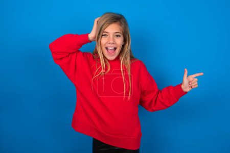 Foto de Sorprendida chica adolescente rubia usando suéter rojo sobre la pared azul apuntando al espacio vacío sosteniendo la mano en la cabeza - Imagen libre de derechos