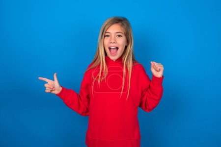 Foto de Rubia adolescente chica vistiendo rojo suéter sobre azul pared puntos en vacío espacio sosteniendo puño arriba, ganador gesto. - Imagen libre de derechos