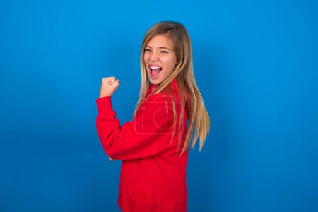 Photo for Profile side view portrait blonde teen girl wearing red sweater over blue wall celebrates victory - Royalty Free Image