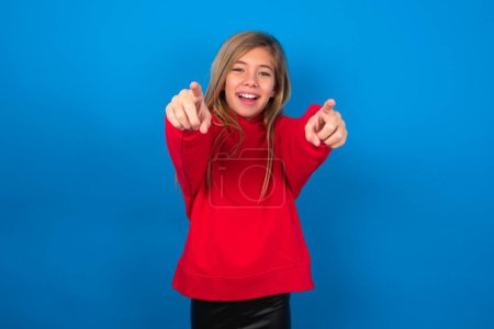 Téléchargez les photos : Blonde adolescent fille portant pull rouge sur le mur bleu joyeux et souriant pointant vers la caméra - en image libre de droit