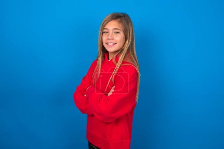 Foto de Retrato de una adolescente rubia con suéter rojo sobre una pared azul de pie con los brazos cruzados y sonriendo - Imagen libre de derechos