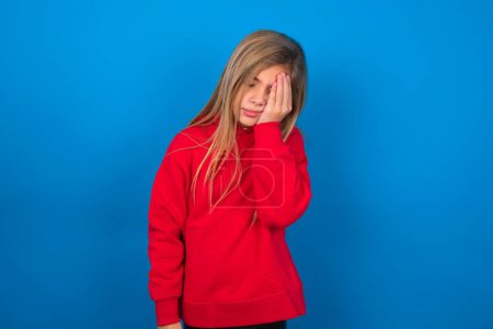 Téléchargez les photos : Blonde adolescente portant un pull rouge sur le mur bleu avec une expression triste couvrant le visage avec les mains tout en pleurant. Concept de dépression. - en image libre de droit