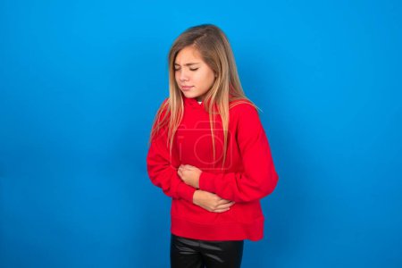 Foto de Rubia adolescente chica usando rojo suéter sobre azul pared tiene dolor de estómago - Imagen libre de derechos