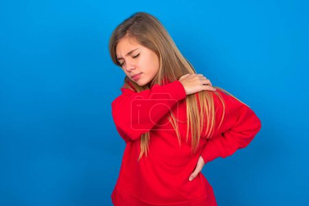 Téléchargez les photos : Blonde adolescent fille portant pull rouge sur le mur bleu a mal au dos - en image libre de droit