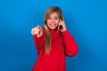 Foto de Positivo rubia adolescente chica usando suéter rojo sobre la pared azul indica directamente a la cámara tiene conversación telefónica sonrisas ampliamente disfruta de hablar largas horas. Te unes a mí - Imagen libre de derechos