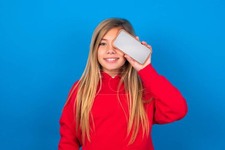 Foto de Rubia adolescente chica usando rojo suéter sobre azul pared celebración de teléfono inteligente moderno que cubre un ojo mientras sonríe - Imagen libre de derechos