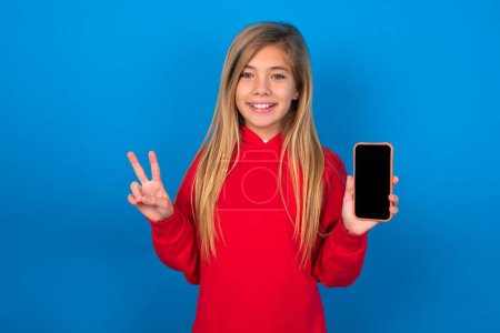 Téléchargez les photos : Blonde adolescent fille portant pull rouge sur le mur bleu tenant dispositif moderne montrant v-signe - en image libre de droit