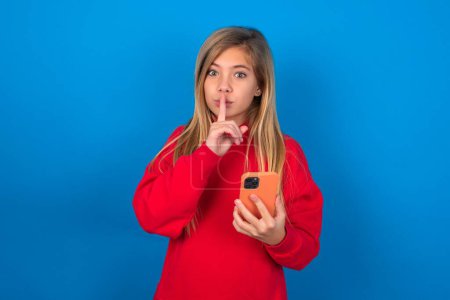 Téléchargez les photos : Blonde adolescent fille portant pull rouge sur le mur bleu tenant gadget moderne demander de ne pas dire de secrets - en image libre de droit