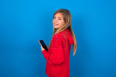 Téléchargez les photos : Vue arrière portrait photo de jeune fille blonde portant un pull rouge sur le mur bleu en utilisant smartphone souriant - en image libre de droit