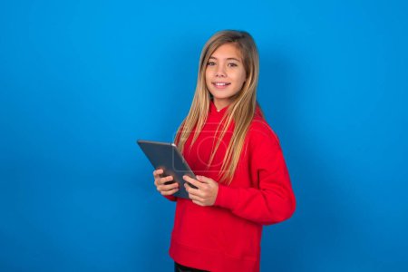 Téléchargez les photos : Photo de jeune fille blonde optimiste portant un pull rouge sur une tablette bleue - en image libre de droit