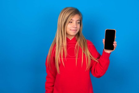 Foto de Foto de asombrada chica adolescente rubia loca usando suéter rojo sobre la pared azul mantenga el concepto de retroalimentación de desagrado del teléfono inteligente - Imagen libre de derechos