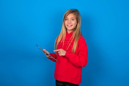 Téléchargez les photos : Photo de joyeuse jeune fille blonde intelligente heureuse portant un pull rouge sur un mur bleu tenir tablette navigation sur Internet - en image libre de droit