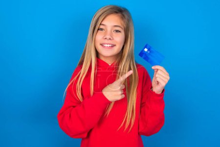 Photo for Photo portrait of blonde teen girl wearing red sweater over blue wall doing purchase with pointing finger credit bank card - Royalty Free Image