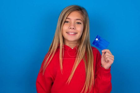 Photo for Photo of happy cheerful smiling positive blonde teen girl wearing red sweater over blue wall recommend credit card - Royalty Free Image