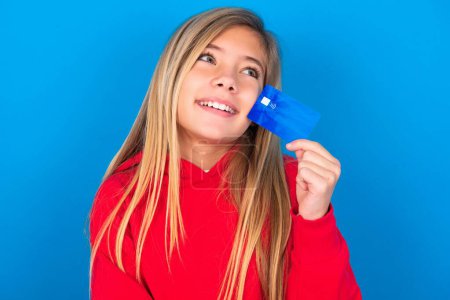 Foto de Hermosa chica adolescente rubia con suéter rojo sobre la pared azul que muestra la tarjeta de crédito y mirando hacia otro lado en el espacio de copia. Pensativo - Imagen libre de derechos