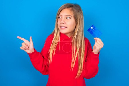 Téléchargez les photos : Curieux sourire blonde adolescent fille portant un pull rouge sur le mur bleu montrant la banque en plastique montrant copyspace doigt - en image libre de droit