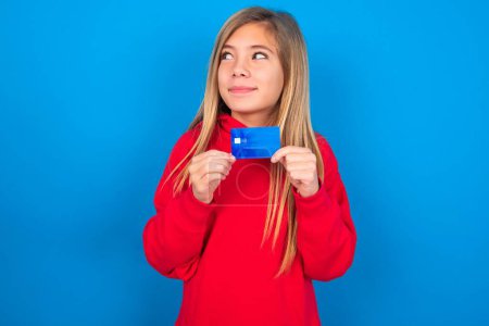 Téléchargez les photos : Photo de gaie jeune fille blonde portant un pull rouge sur le mur bleu tenir carte de débit regarder espace vide - en image libre de droit