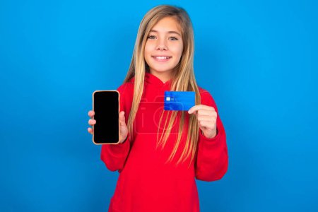 Téléchargez les photos : Photo d'adorable jeune fille blonde portant un pull rouge sur le mur bleu tenant la carte de crédit et Smartphone. Réservé aux achats en ligne - en image libre de droit