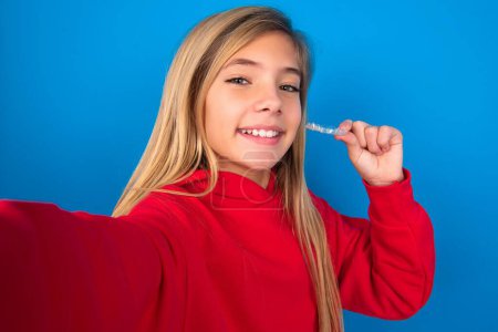 Téléchargez les photos : Blonde adolescente portant un pull rouge sur le mur bleu font selfie tenant un aligneur de bretelles invisibles, recommandant ce nouveau traitement. Concept de soins dentaires. - en image libre de droit