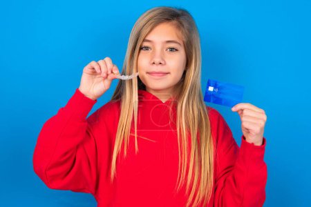 Photo for Blonde teen girl wearing red sweater over blue wall holding a credit card and invisible aligner. Ready to finance treatment - Royalty Free Image