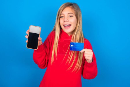 Foto de Rubia adolescente chica vistiendo suéter rojo sobre la pared azul abrió cuenta bancaria, celebración de teléfono inteligente y tarjeta de crédito, sonriendo, recomienda el uso de la aplicación de compras en línea - Imagen libre de derechos