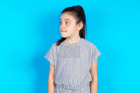 Photo for Amazed Beautiful kid girl wearing striped dress over blue background bitting lip and looking tricky to empty space. - Royalty Free Image