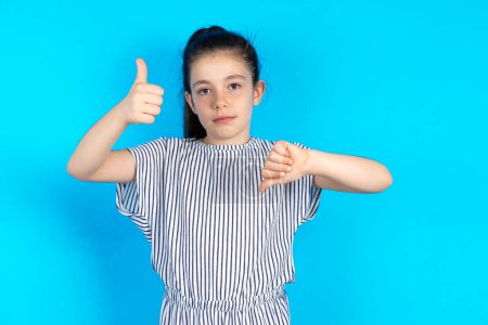 Photo for Beautiful kid girl wearing striped dress over blue background showing thumbs up and thumbs down, difficult choose concept - Royalty Free Image