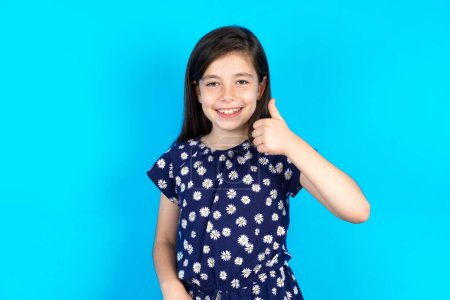 Photo for Beautiful kid girl wearing dress over blue background doing happy thumbs up gesture with hand. Approving expression looking at the camera showing success. - Royalty Free Image