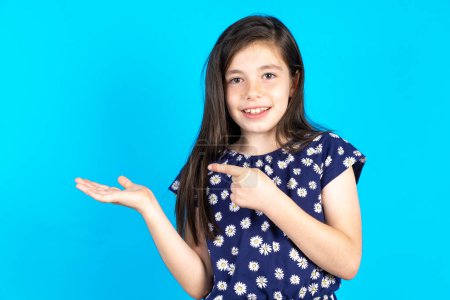 Photo for Beautiful kid girl wearing dress over blue background pointing and holding hand showing adverts - Royalty Free Image