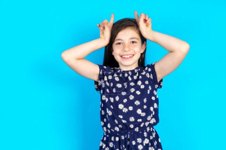 Foto de Divertido Hermosa niña con vestido floral sobre fondo azul muestra cuernos, dedos en el gesto de la cabeza, posando tonto y lindo - Imagen libre de derechos
