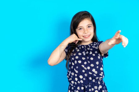 Téléchargez les photos : Belle fille enfant portant une robe sur fond bleu pointant vers la caméra avec un sourire satisfait, confiant et amical, vous choisissant - en image libre de droit