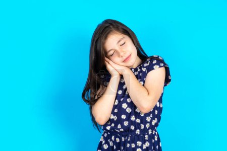 Photo for Beautiful brunette caucasian kid girl wearing dress over blue background with closed eyes leaning on palms making sleeping gesture. - Royalty Free Image