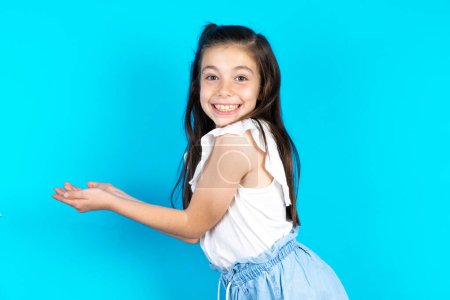 Téléchargez les photos : Belle fille enfant caucasien debout sur fond bleu studio pointant de côté avec les mains ouvertes paumes montrant l'espace de copie, présentant la publicité souriant heureux excité - en image libre de droit