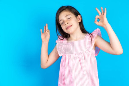 Téléchargez les photos : Belle petite fille portant une robe rose sur fond bleu se détendre et sourire les yeux fermés en faisant geste de méditation avec les doigts. Concept de yoga. - en image libre de droit