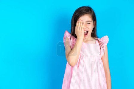 Foto de Hermosa niña con vestido rosa sobre fondo azul bostezo cansado cubriendo media cara, ojo y boca con la mano. La cara duele de dolor. - Imagen libre de derechos