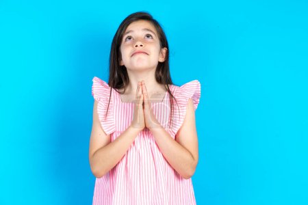 Foto de Hermosa niña con vestido rosa sobre fondo azul mendigando y rezando con las manos junto con la expresión de esperanza en la cara muy emocional y preocupado. Por favor Dios - Imagen libre de derechos