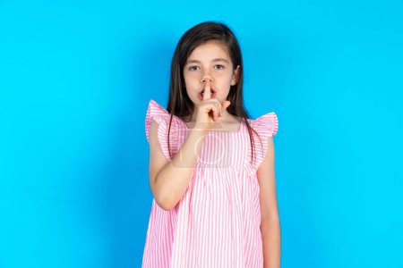 Photo for Beautiful kid girl wearing pink dress over blue background makes silence gesture, keeps finger over lips. Silence and secret concept. - Royalty Free Image