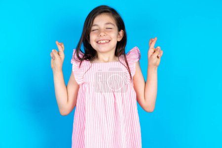 Photo for Beautiful kid girl wearing pink dress over blue background gesturing finger crossed smiling with hope and eyes closed. Luck and superstitious concept. - Royalty Free Image