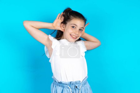 Téléchargez les photos : Belle gamine caucasienne debout sur fond bleu studio Essayant d'entendre les deux mains sur le geste de l'oreille, curieuse de commérages. Problème auditif, sourd - en image libre de droit