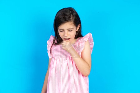 Foto de Hermosa niña con vestido rosa sobre fondo azul sensación de malestar y tos como síntoma de resfriado o bronquitis. Concepto sanitario. - Imagen libre de derechos
