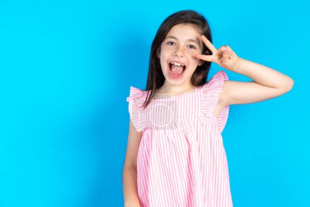 Photo for Beautiful kid girl wearing pink dress over blue background Doing peace symbol with fingers over face, smiling cheerful showing victory - Royalty Free Image