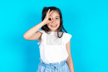Photo for Beautiful caucasian kid girl standing over blue studio background doing ok gesture with hand smiling, eye looking through fingers with happy face. - Royalty Free Image