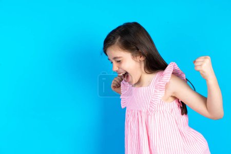 Téléchargez les photos : Belle fille enfant portant une robe rose sur fond bleu très heureux et excité faisant geste gagnant avec les bras levés, souriant et criant pour le succès. Concept de célébration. - en image libre de droit
