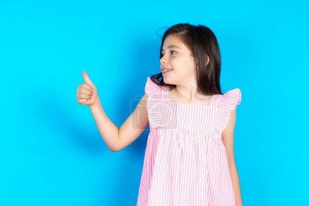 Photo for Kid Looking proud, smiling doing thumbs up gesture to the side. Good job! Beautiful caucasian little girl posing over blue studio background - Royalty Free Image
