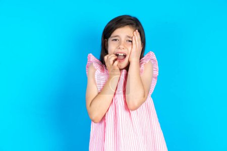 Photo for Doleful desperate crying kid, looks stressfully, frowns face, feels lonely and anxious. Beautiful caucasian little girl posing over blue studio background - Royalty Free Image