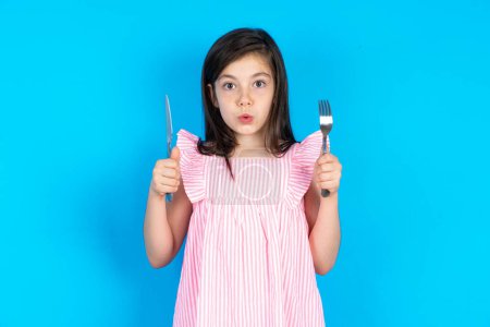 Photo for Hungry kid holding in hand fork knife want tasty yummy pizza pie. Beautiful caucasian little girl posing over blue studio background - Royalty Free Image