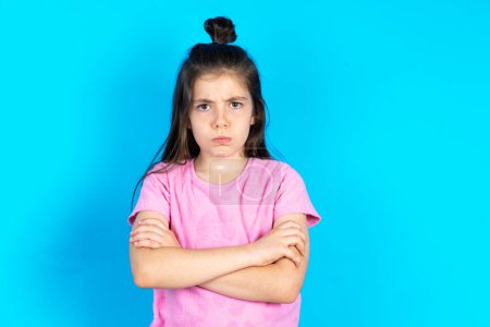 Photo for Gloomy dissatisfied kid looks with miserable expression at camera from under forehead, makes unhappy grimace. Beautiful caucasian little girl posing over blue studio background - Royalty Free Image