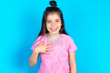 Photo for Kid smiles broadly,cannot believe her eyes, expresses good emotions and surprisement. Beautiful caucasian little girl posing over blue studio background - Royalty Free Image