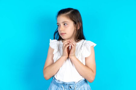 Photo for Kid steepling fingers and looking mysterious aside, has great evil plan on mind. Beautiful caucasian little girl posing over blue studio background - Royalty Free Image