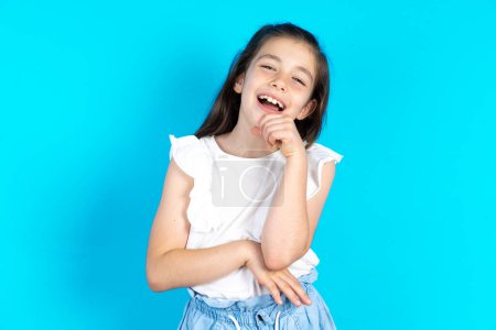 Photo for Kid laughs happily, keeps hand on chin, expresses positive emotions. Beautiful caucasian little girl posing over blue studio background - Royalty Free Image
